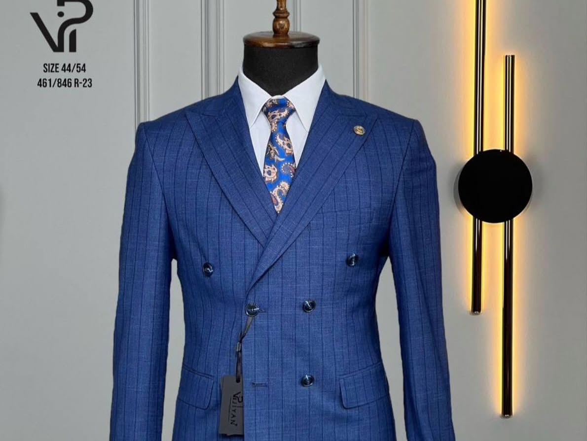 A blue double-breasted pinstriped suit with a patterned tie is shown on a headless mannequin, lit by warm vertical lights.