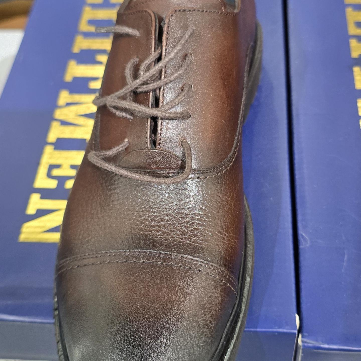 Close-up photograph of a brown leather lace-up dress shoe on a blue shoebox, showing the toe and lacing from above in bright indoor lighting.