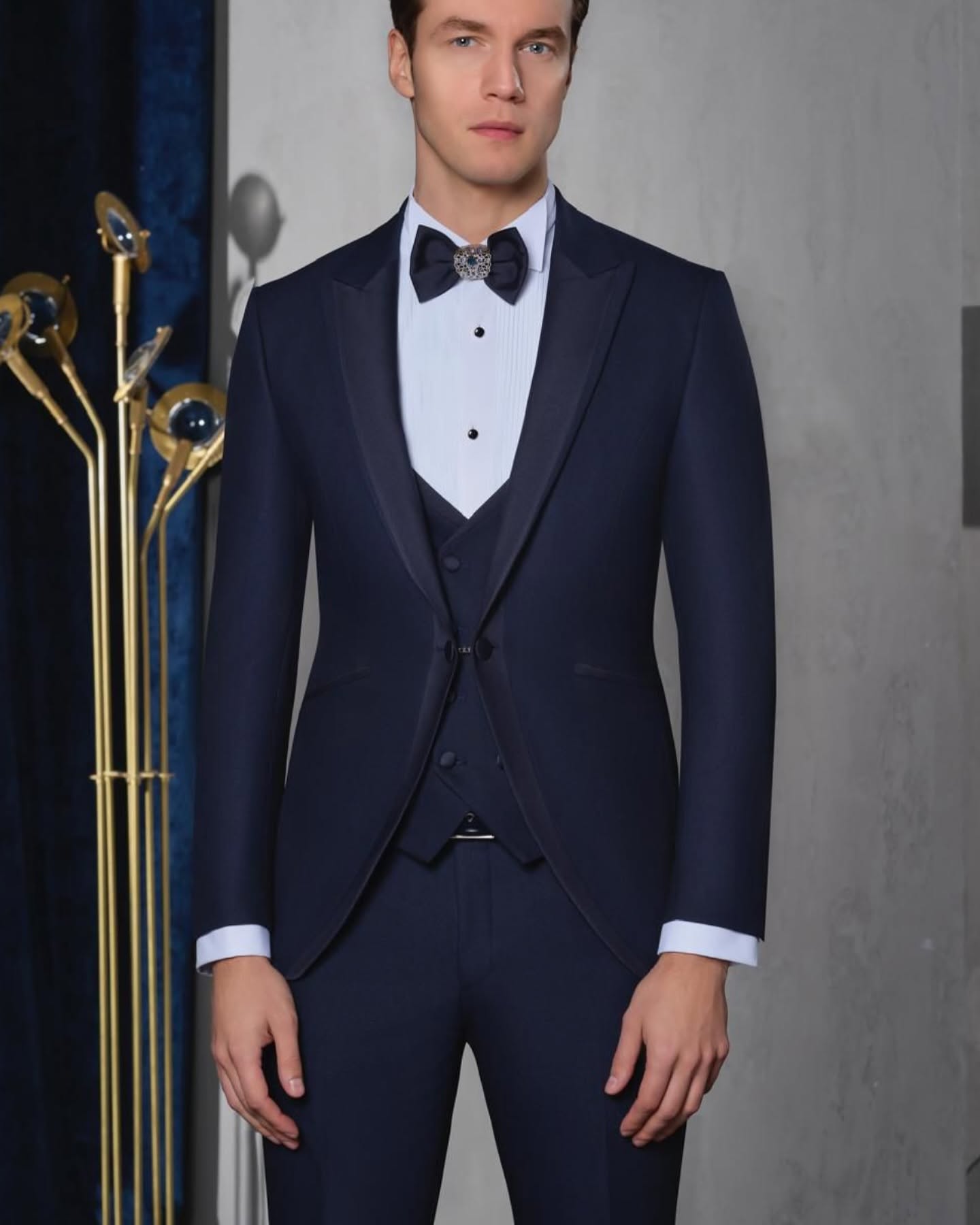 Studio-style portrait of a man wearing a navy tuxedo, framed from head to knees against a dark interior backdrop.