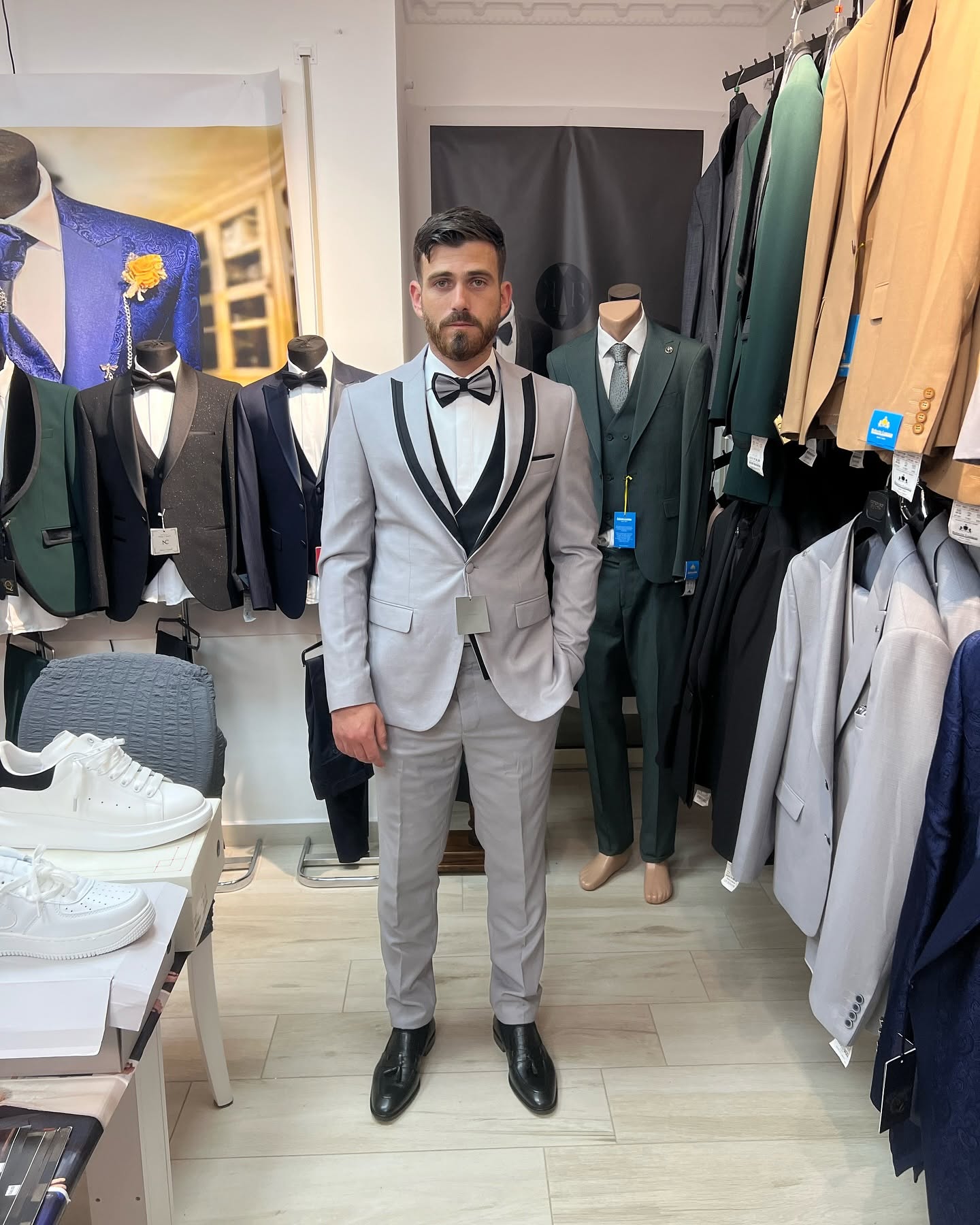Full-body view of a man wearing a light gray tuxedo in a showroom, framed among displayed suits and racks.