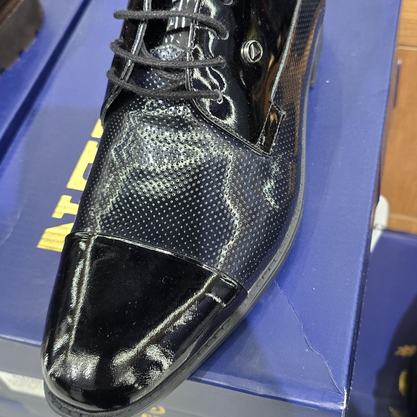 Close-up photograph of a black leather lace-up dress shoe with perforated detailing on a blue shoebox, brightly lit indoors.