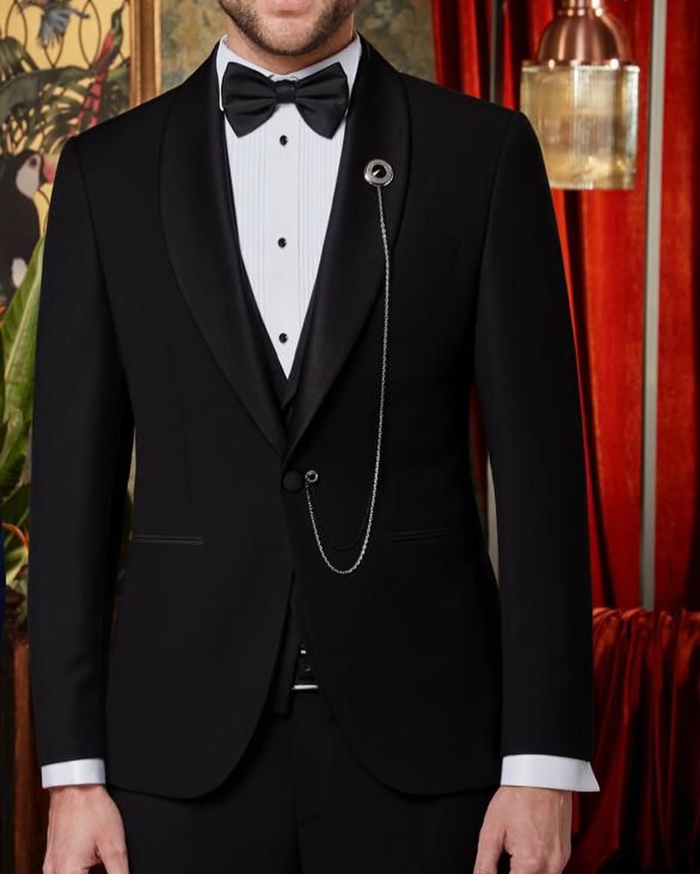 Cropped studio portrait of a man wearing a black tuxedo with a bow tie and chain accessory, warmly lit against a red-toned interior.
