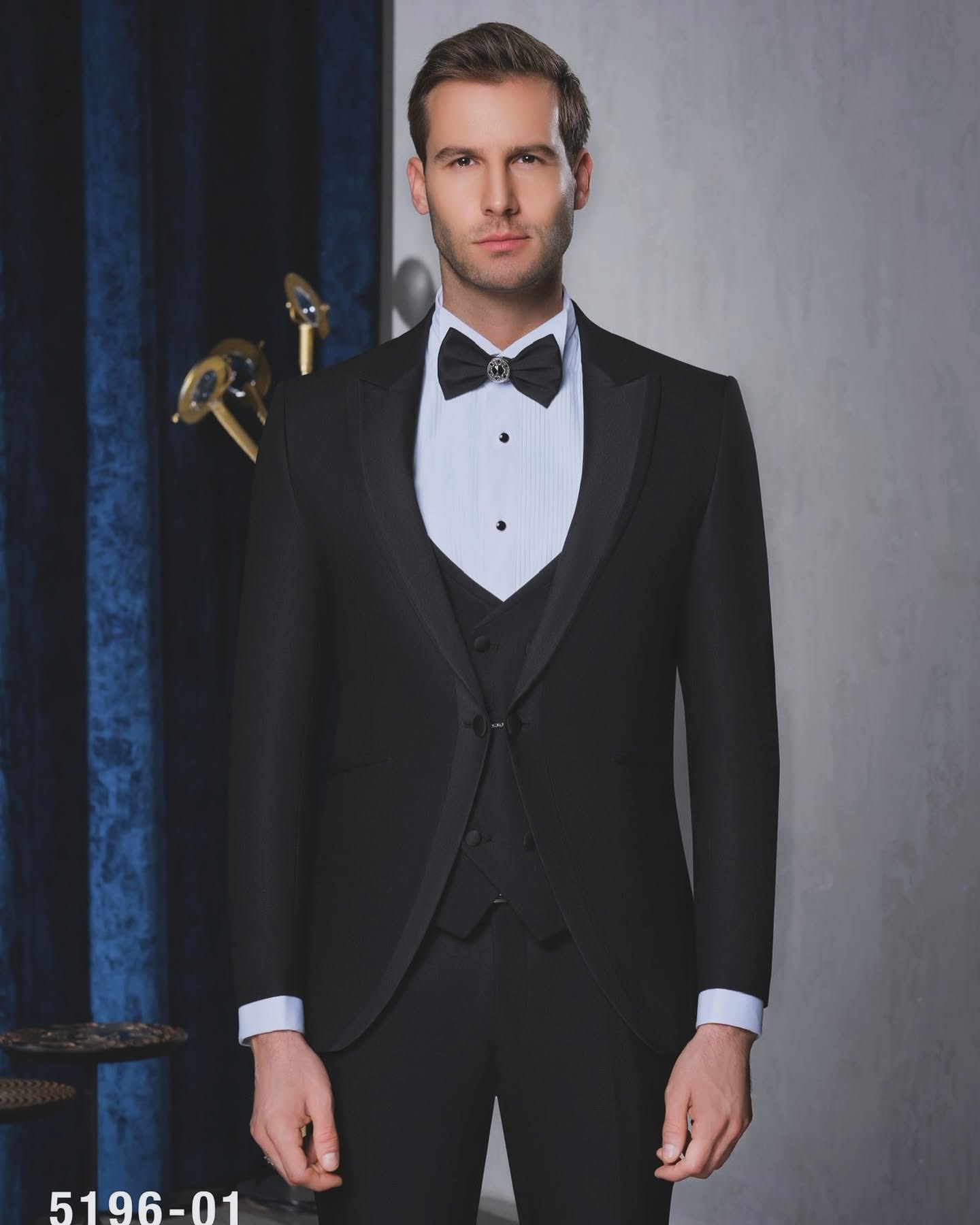 A man poses from the knees up in a black tuxedo and bow tie against a dark, softly lit background.