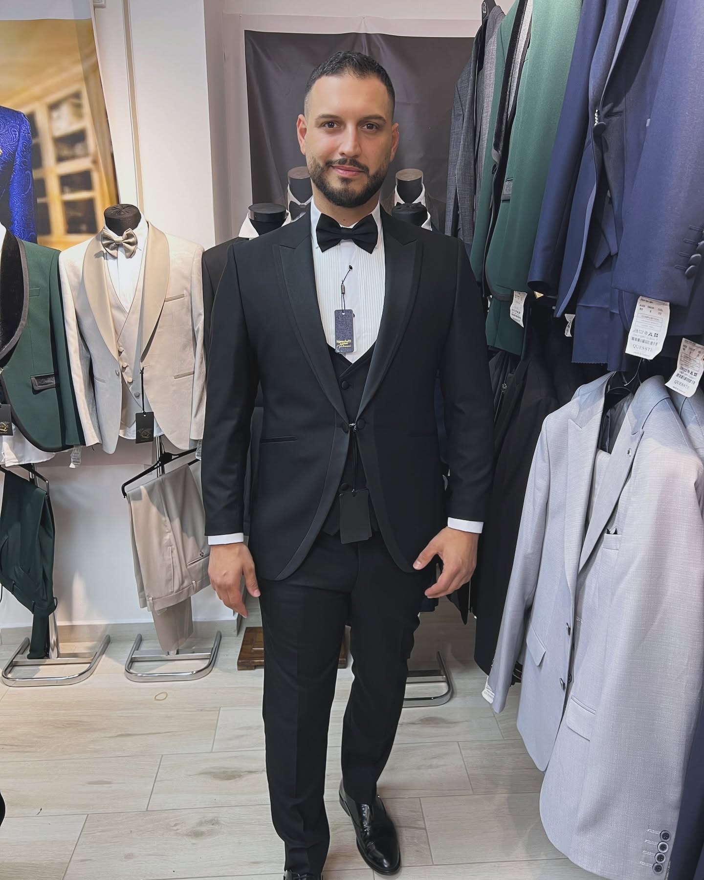 A man stands wearing a black tuxedo and bow tie, framed by hanging suits and mannequins in bright indoor lighting.