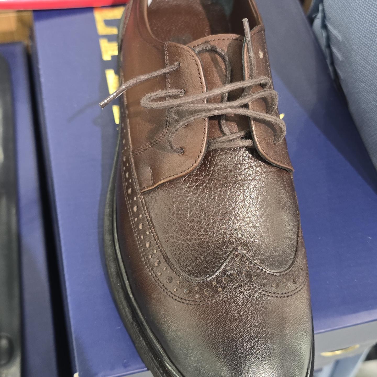 Close-up photograph of a brown leather lace-up dress shoe resting on a blue shoebox, viewed from above under bright indoor lighting.